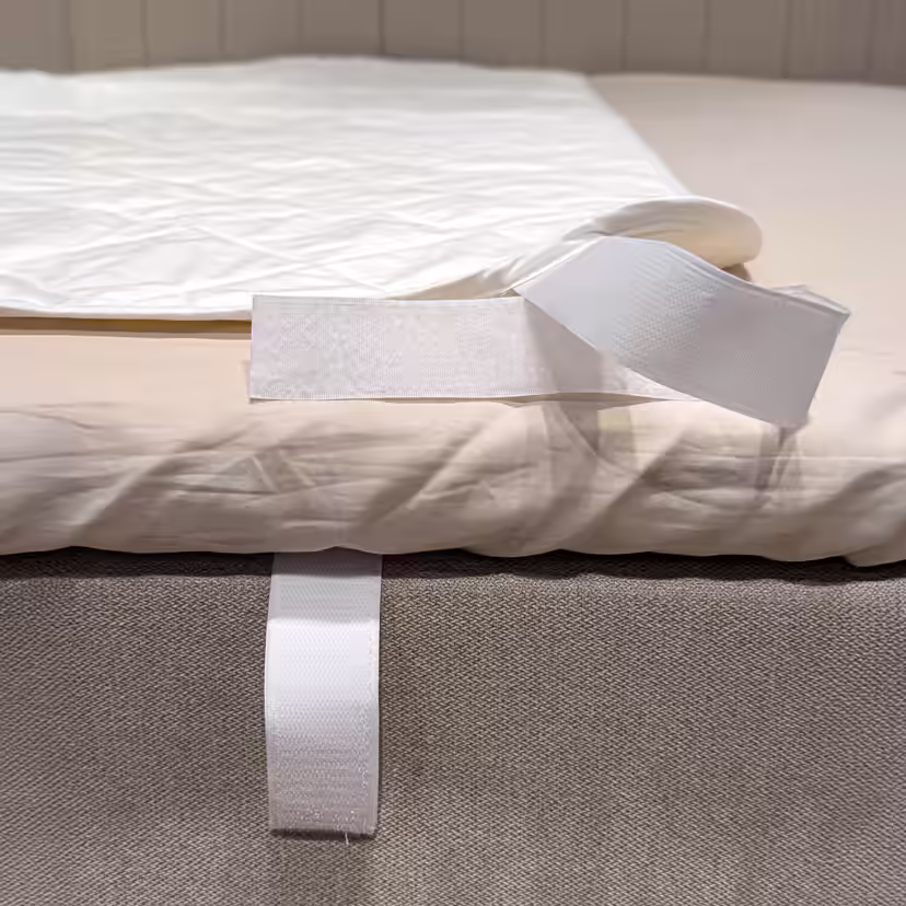 Odchylony narożnik białego pikowanego ochraniacza na materac, odsłaniający pasek z rzepem biegnący wokół krawędzi łóżka